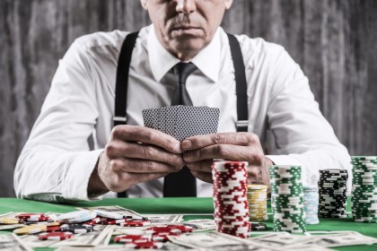 Poker player. Close-up of serious senior man in shirt and suspenders sitting at the poker table and holding cards  with money and  gambling chips laying all around him